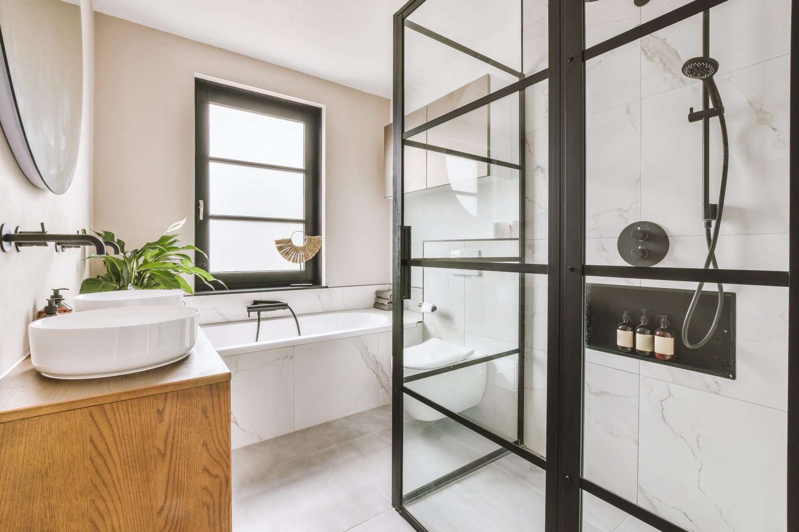 Shower faucets attached to tiled wall near glass partition, toilet, sink and ornamental curtail in washroom at home